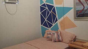 A small, neatly made bed with a pink bedspread and two pillows, a wooden nightstand with a lamp, and a wall with a geometric pattern and a framed painting of a flamingo.