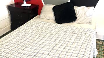 A bed with a checkered bedspread, a black pillow, and a white pillow. There is a nightstand with a fan and a red wall.