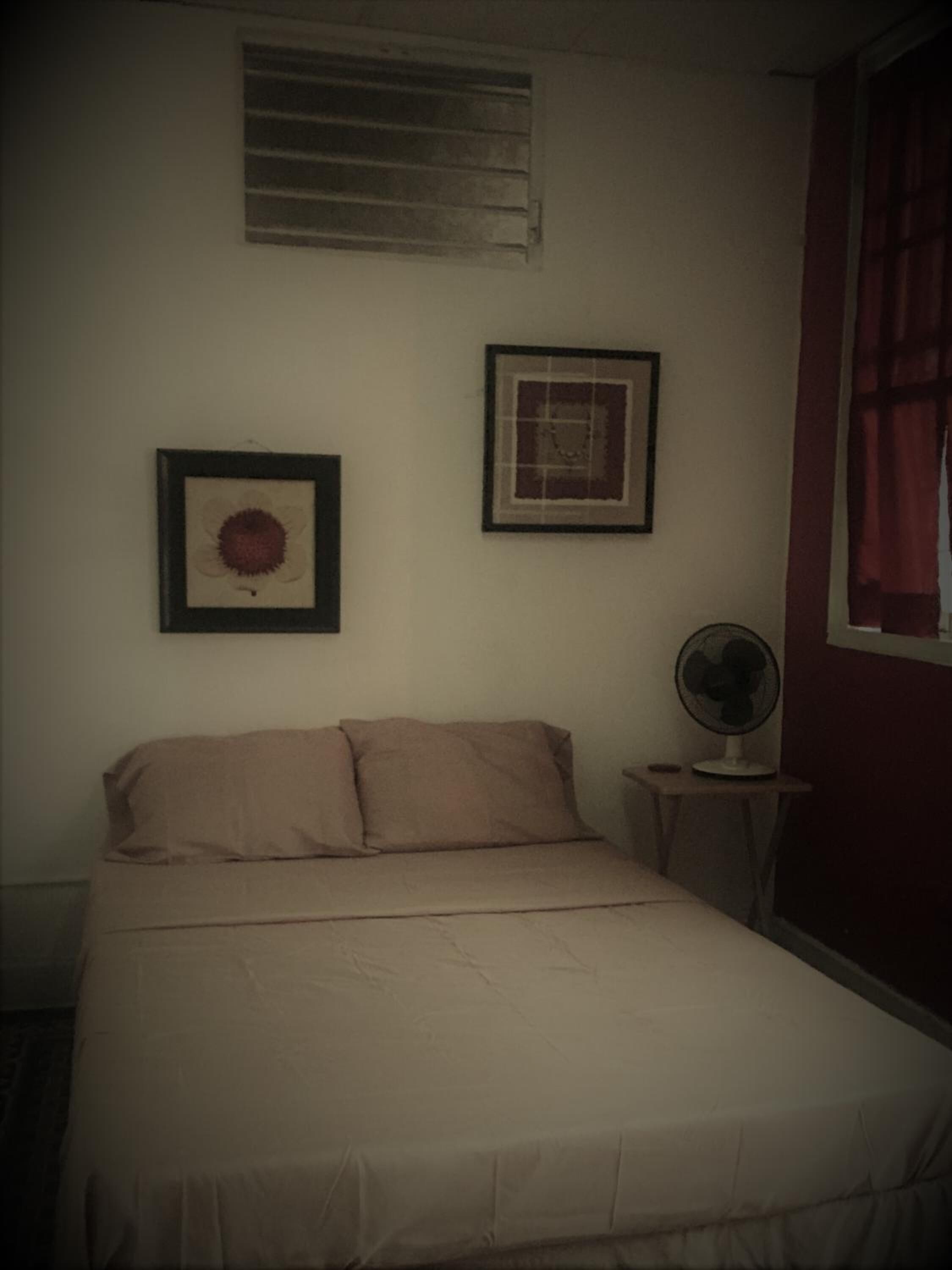 A bedroom with a bed, a wall-mounted fan, and two framed pictures.