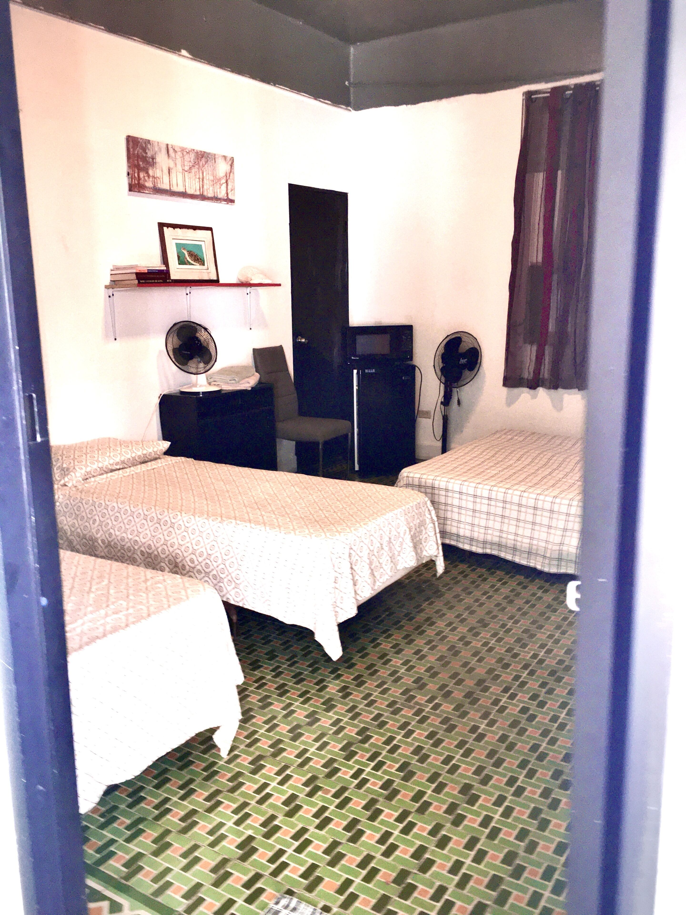 A small, compact room with two single beds, a desk with a computer, a chair, a shelf with books, and a wall-mounted fan.