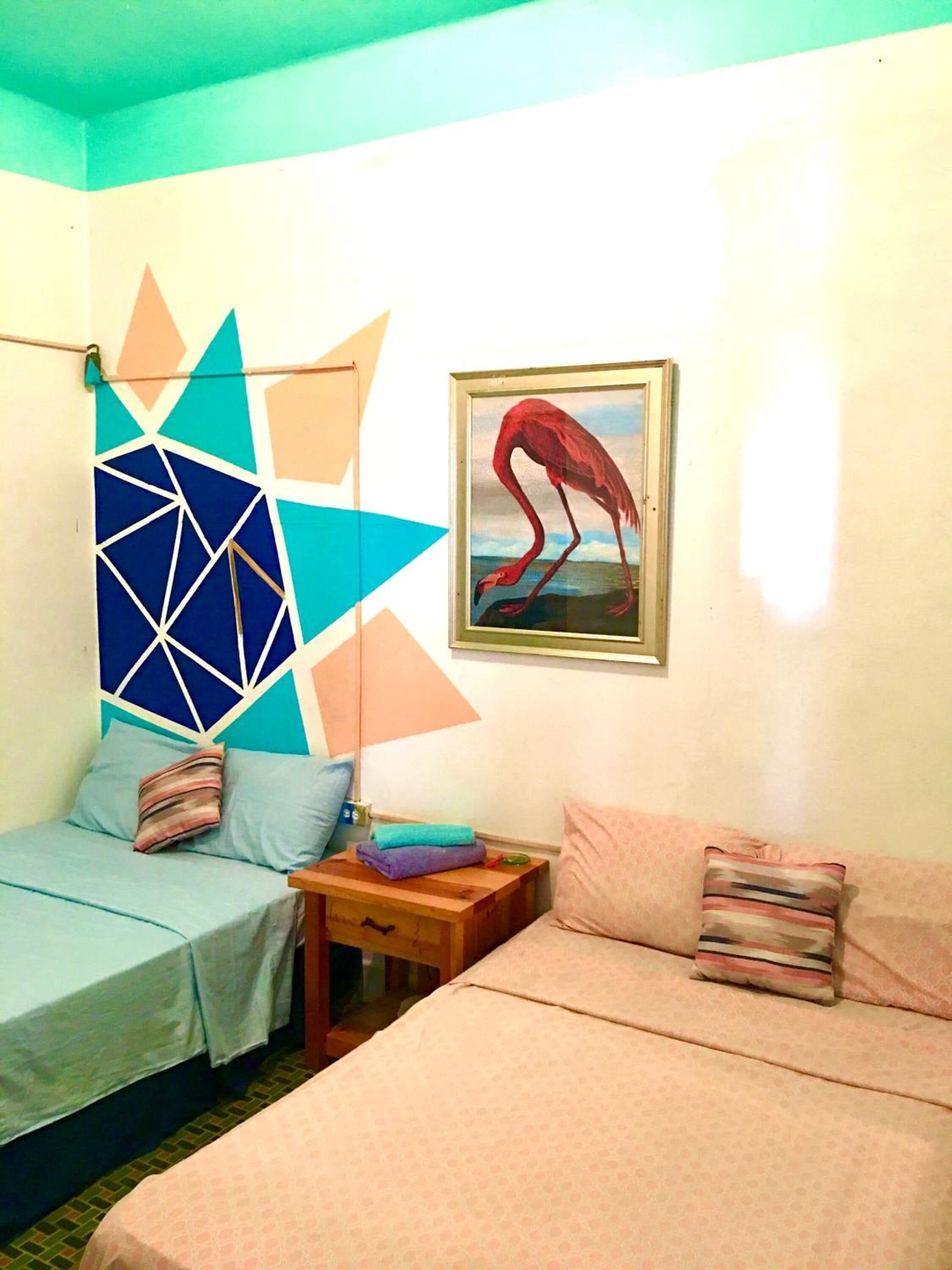Una habitación con dos camas, una con cabecera de estampado geométrico y la otra con cabecera lisa. Hay un cuadro con una imagen de un flamenco en la pared.