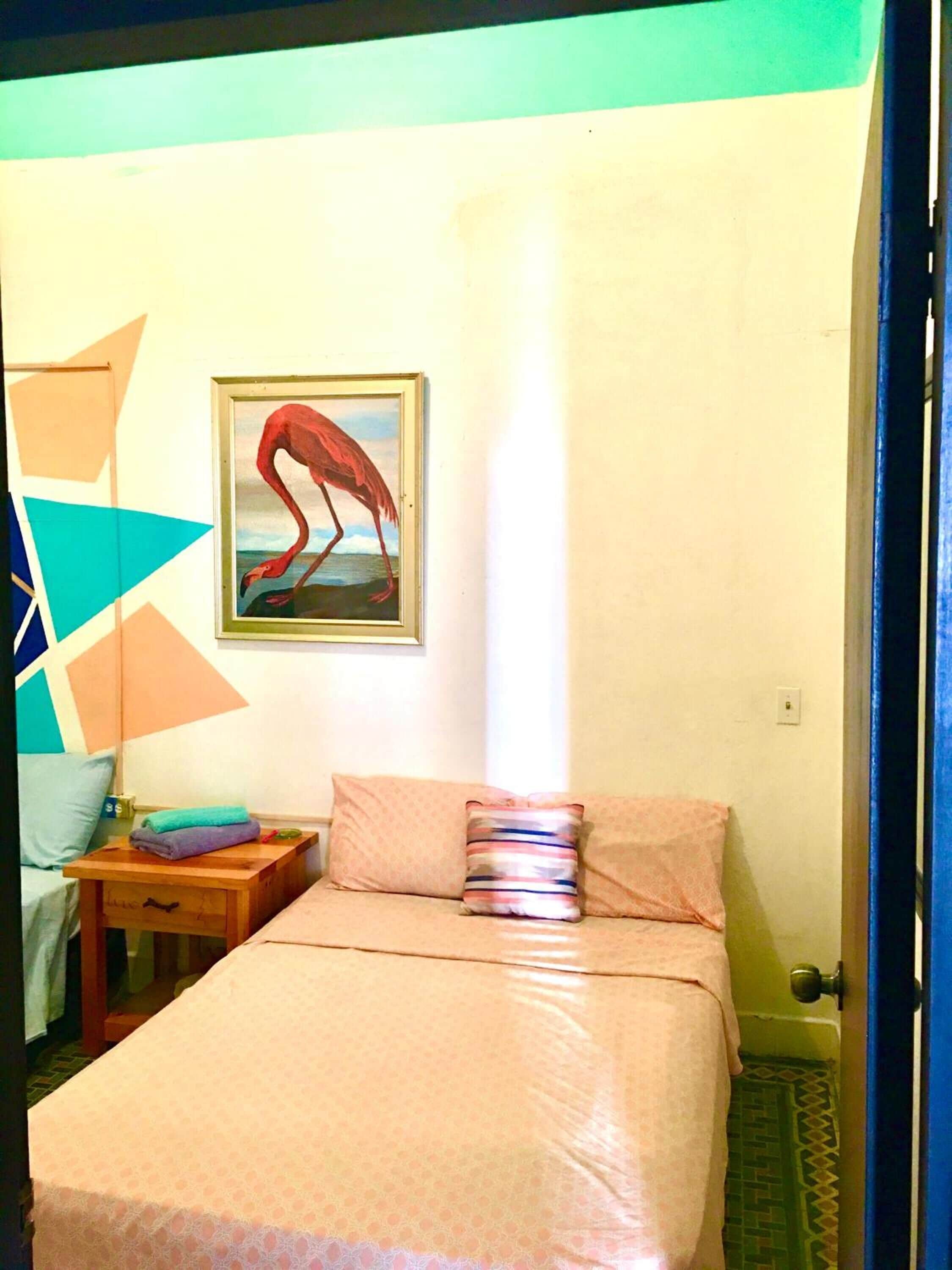 A bedroom with a bed, pillows, a nightstand, a painting of a flamingo, and a geometric wall decoration.
