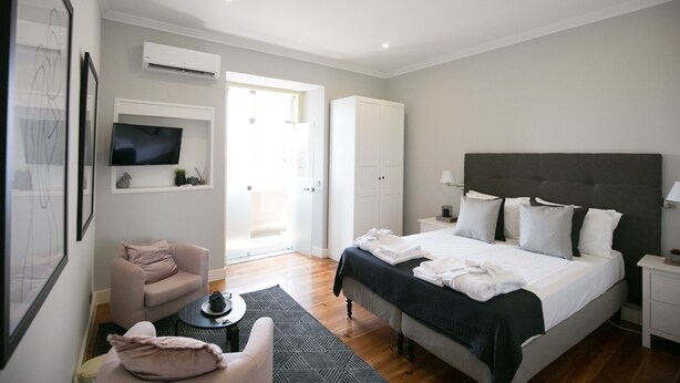 A modern bedroom with a large bed, a TV mounted on the wall, a wardrobe, and a seating area with a chair and a small table. - Bhuiyan Family Host (Lisbon)