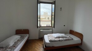 A small, clean room with two single beds, a window with a view of buildings, and a wall-mounted air conditioning unit. - Villa Luzzatti Hostel (Rome)