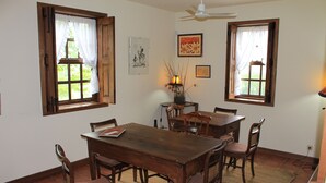 A dining room with a wooden table and chairs, two windows with curtains, a ceiling fan, and a lamp on a side table. - Quinta Do Pomarinho (Arouca)