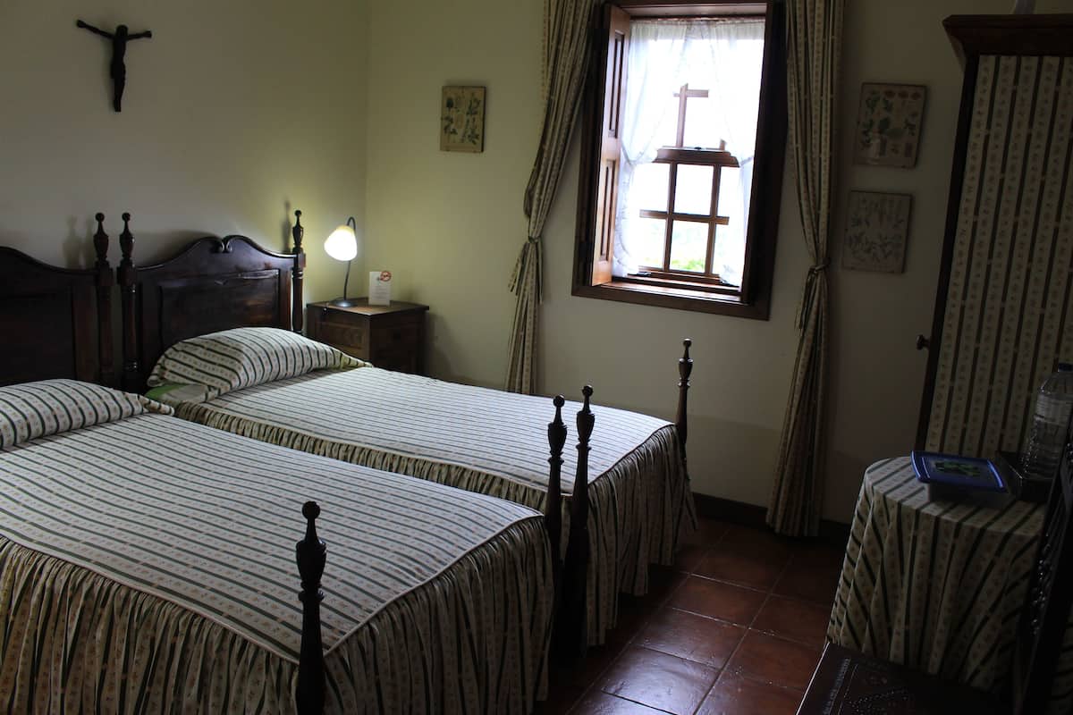 A bedroom with two beds, a nightstand, a window with curtains, and a small table with a lamp.