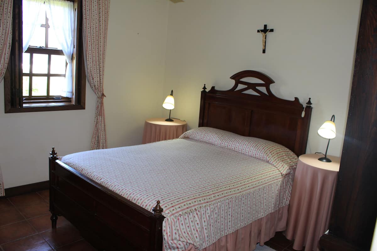 A bedroom with a wooden bed, two bedside tables with lamps, a window with curtains, and a cross on the wall.