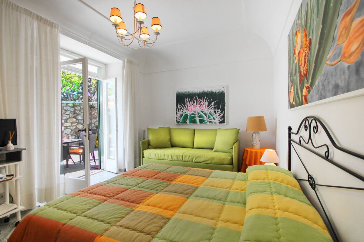 Ein Schlafzimmer mit Bett, einem grünen Sofa, einem Gemälde und Blick ins Freie.