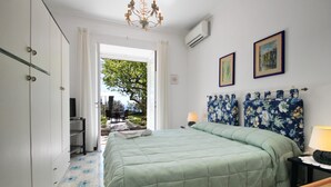 Un dormitorio con una cama, un escritorio y una silla. Se ve una ventana con vistas a un árbol y un edificio.