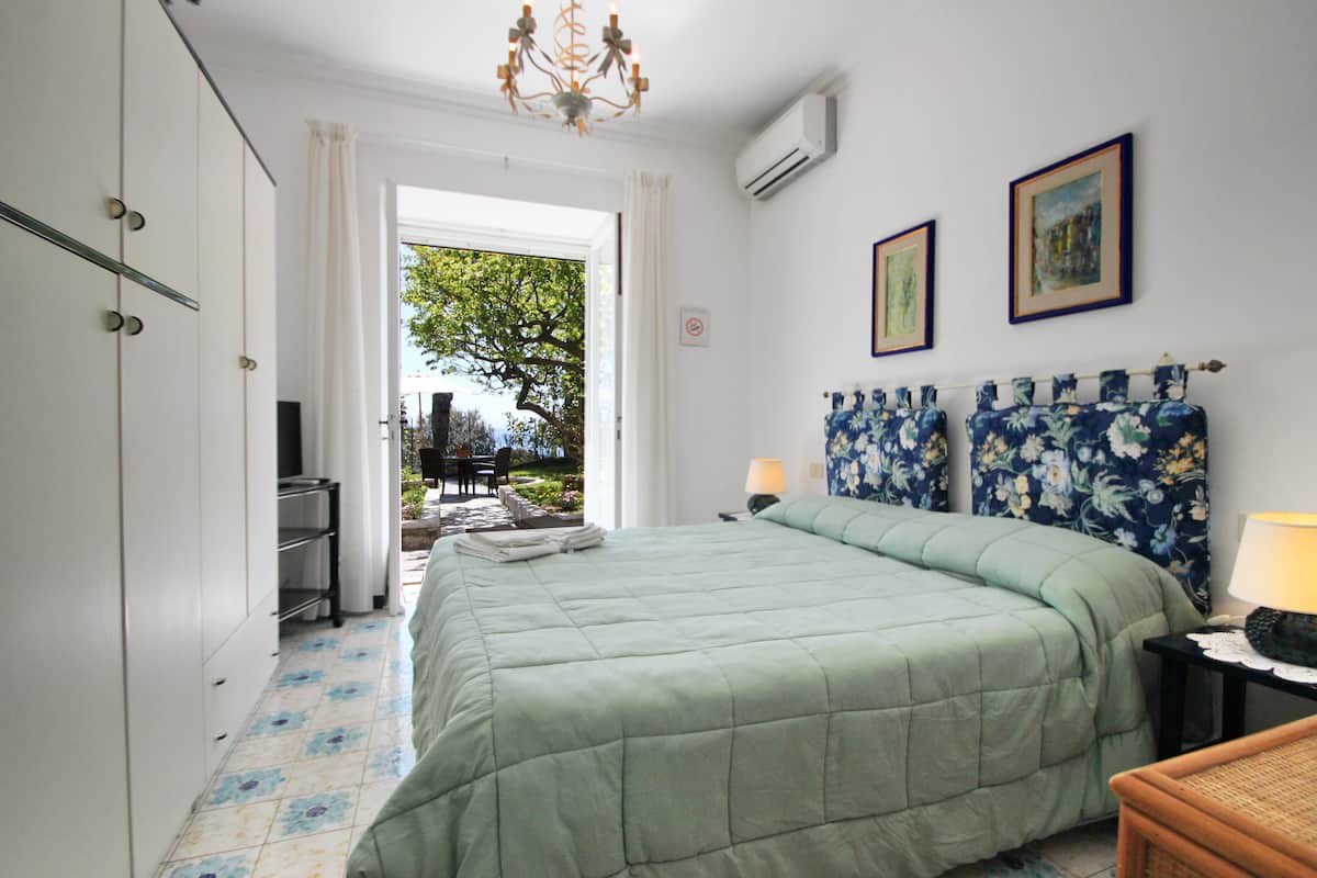 Ein Schlafzimmer mit einem Bett, einem Schreibtisch und einem Stuhl. Ein Fenster bietet Blick auf einen Baum und ein Gebäude.