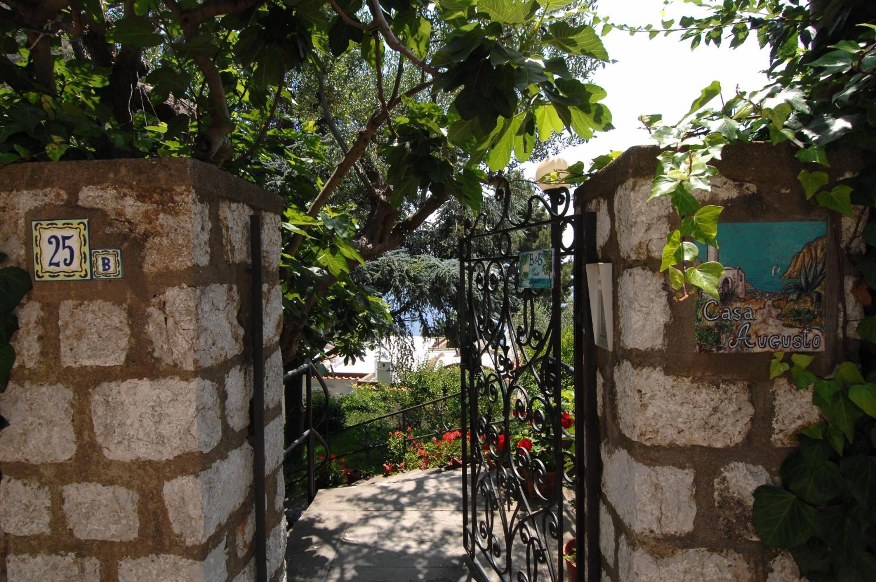 A stone gate with a metal gate, surrounded by greenery and a house with a colorful mural.
