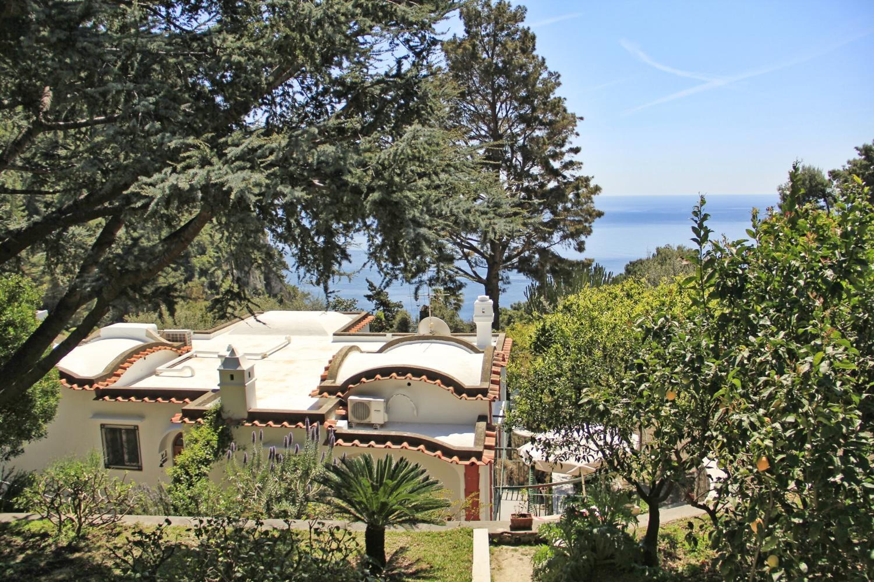 A Mediterranean-style villa with a terracotta roof, surrounded by lush greenery and overlooking a clear blue sea.