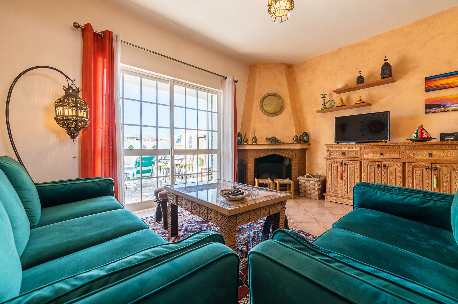 Un salon avec deux canapés de couleur turquoise, une table basse en bois, une cheminée, un téléviseur et un balcon agrémenté de rideaux rouges.