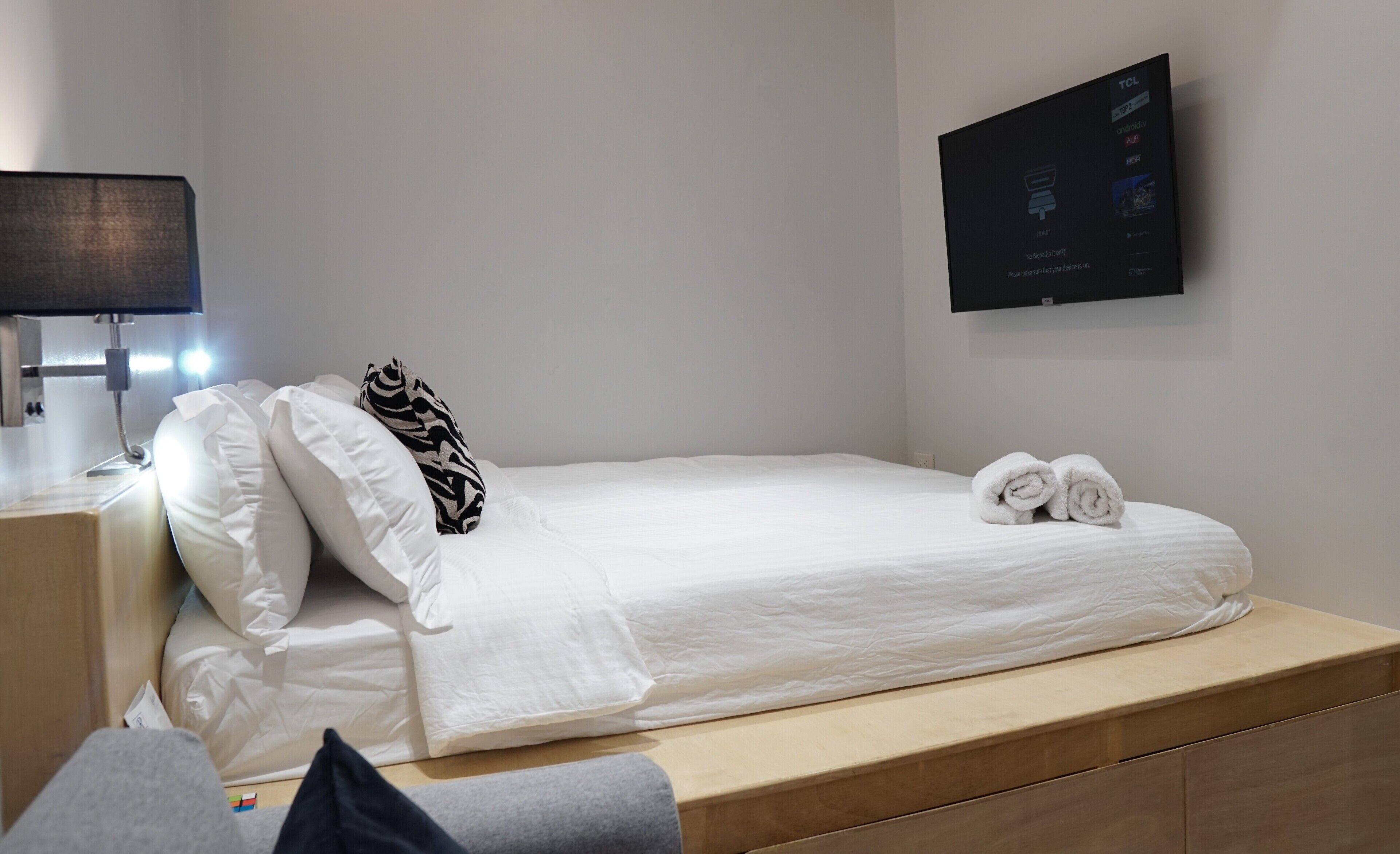 Un lit bien fait, avec du linge de lit blanc et un coussin à motifs zébrés, une petite table de chevet avec une lampe, et un téléviseur fixé au mur qui affiche un menu.