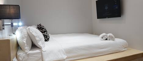Una cama bien hecha con ropa blanca y un cojín estampado con diseño de cebra, una mesita de noche pequeña con una lámpara, y un televisor en la pared mostrando un menú.
