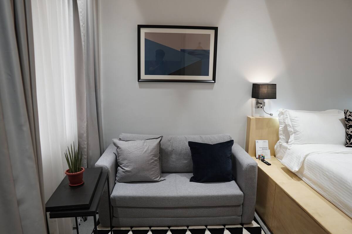 Una habitación de hotel moderna con un sofá gris, una mesita con una planta en maceta, una obra de arte enmarcada en la pared, una lámpara de noche y una cama bien hecha.