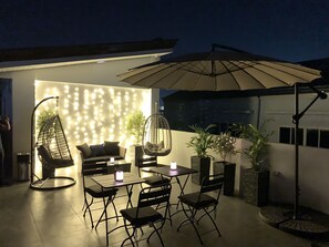 Un espacio al aire libre con patio amoblado, que incluye un sofá, sillas, una mesa y una silla colgante. Hay plantas en macetas y un paraguas que da sombra.