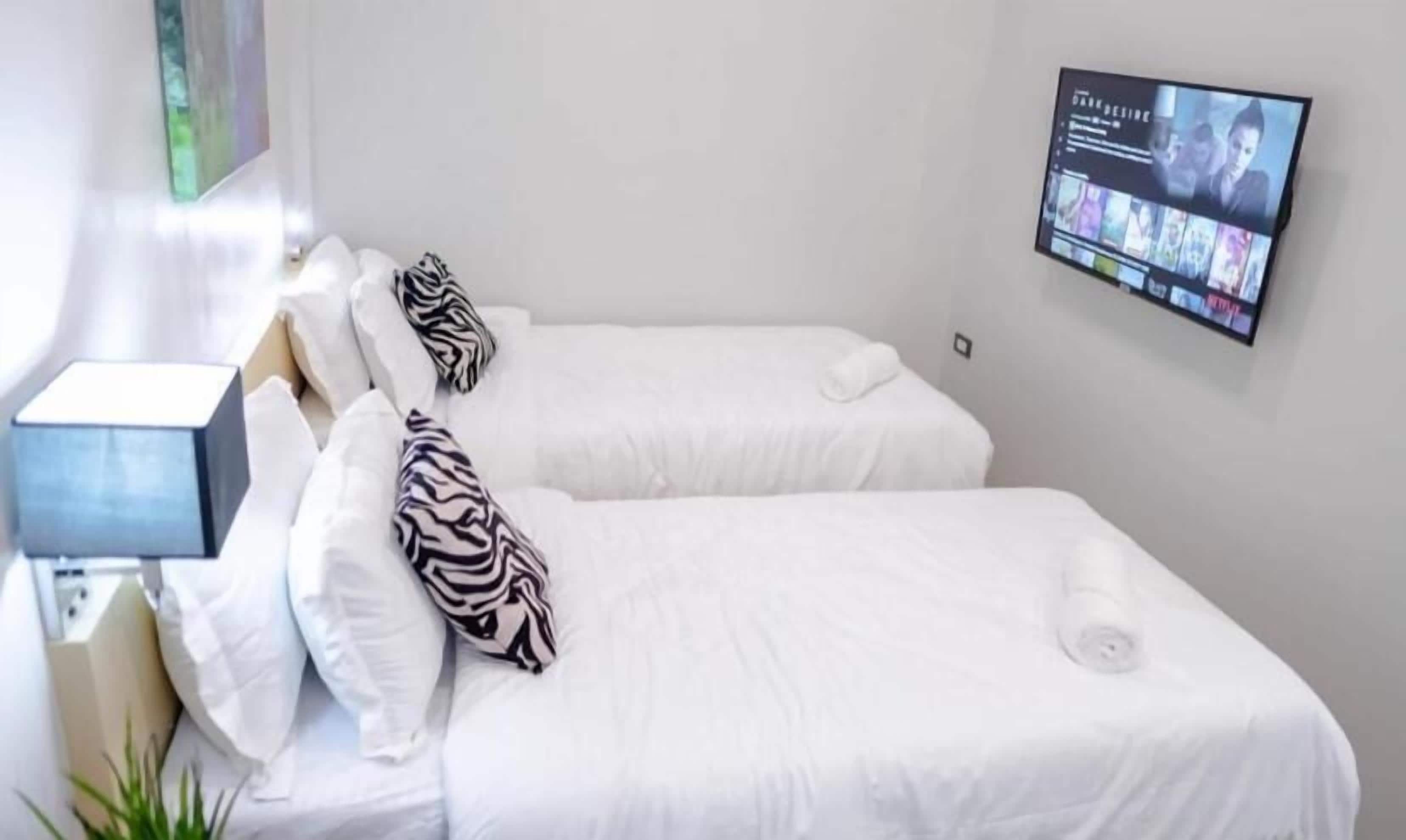 A hotel room with two beds, each with white bedding and zebra-patterned pillows. A flat-screen TV is mounted on the wall above the beds, displaying a movie.