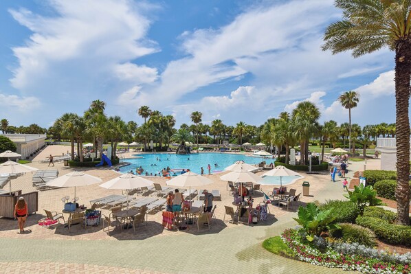 A resort with a swimming pool, lounge chairs, umbrellas, and palm trees.