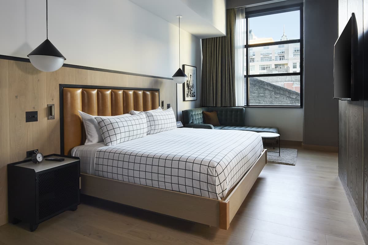 Une chambre d’hôtel moderne dotée d’un grand lit, de tables de chevet, d’un coin salon et offrant une vue sur la ville grâce à une grande fenêtre.