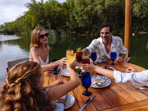 A group of people dining outdoors at a wooden table with a view of a river and trees. - Fairmont Heritage Place Mayakoba (Playa del Carmen)