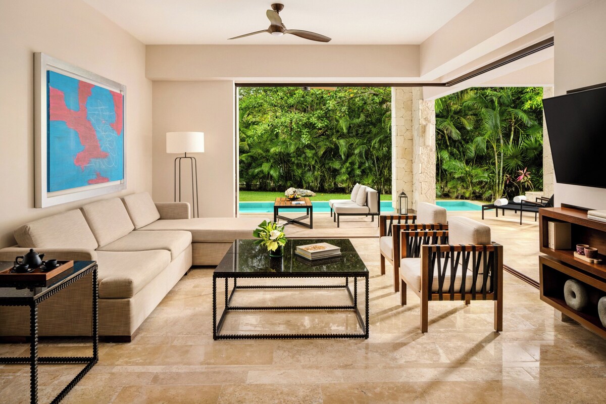 Ein modernes Wohnzimmer mit einer Couch, Couchtisch und Sesseln, einem Fernseher und Blick auf einen Swimmingpool und Grünflächen.