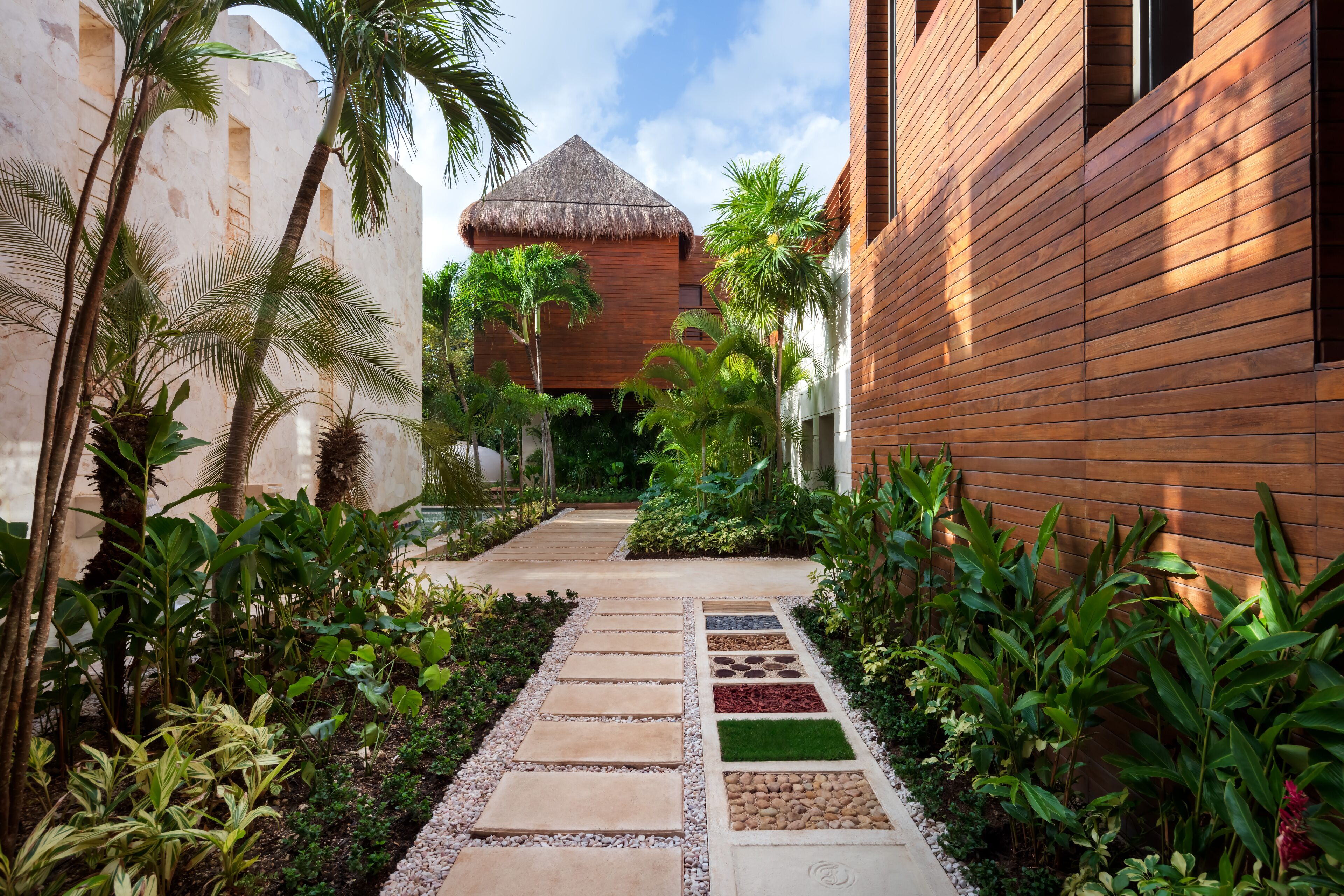 Een tropisch resort met een rieten gebouw, een houten constructie en een verhard pad omzoomd met tropische planten.