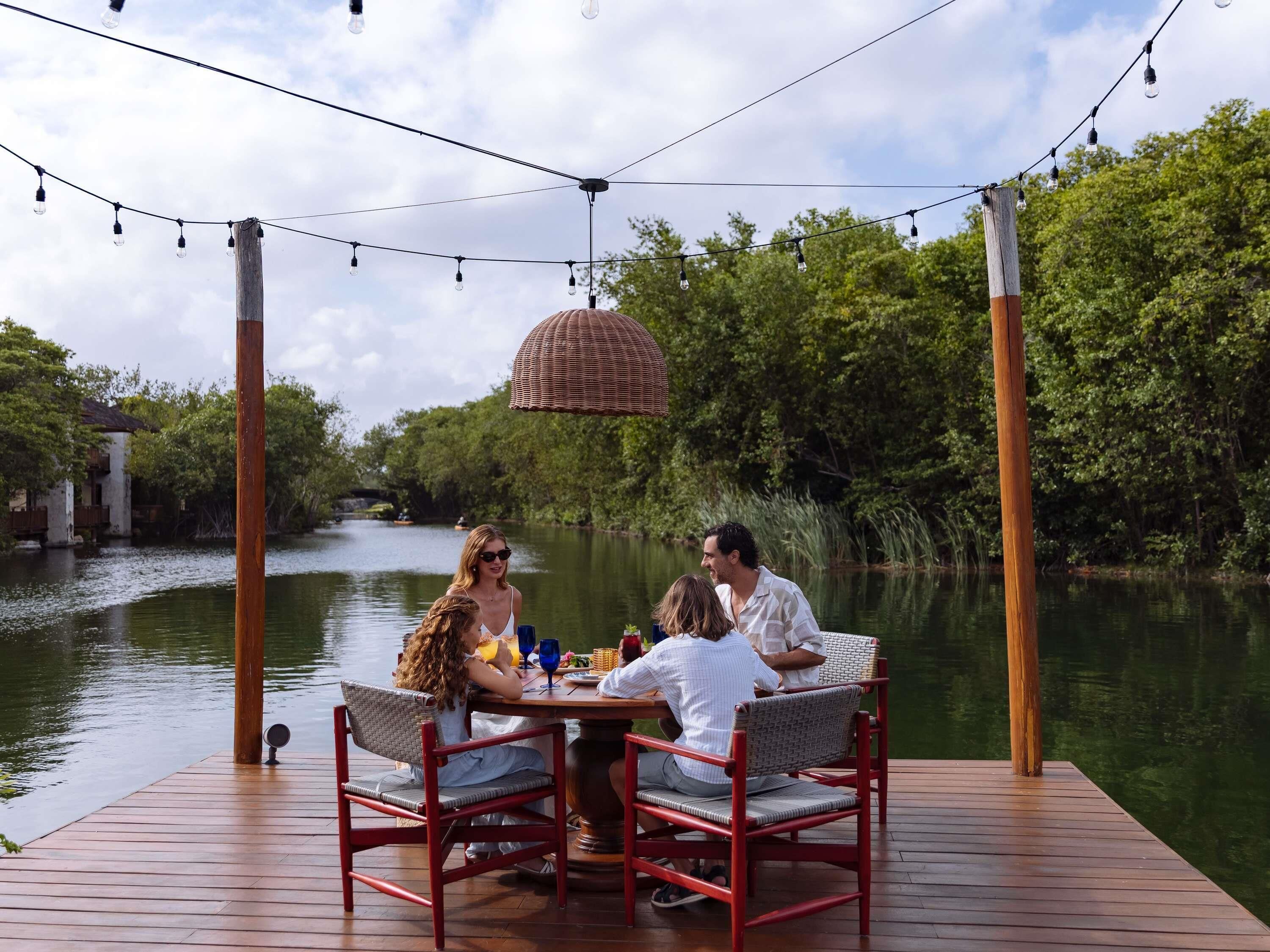 Een houten terras met een tafel en stoelen, uitkijkend op een rivier met begroeiing en een gebouw op de achtergrond.