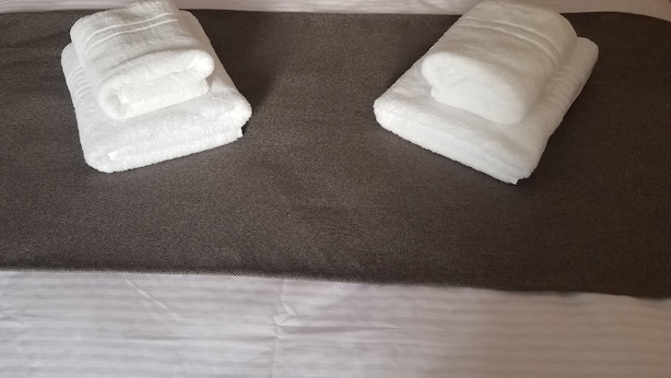 Una cama bien hecha con sábanas blancas y dos toallas dobladas sobre un edredón marrón.