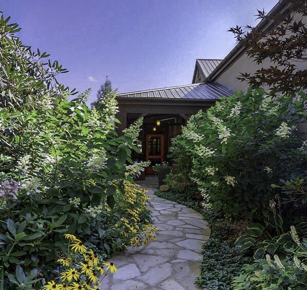 A stone pathway leading to a building with a metal roof, surrounded by lush greenery and flowering plants.