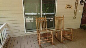 Deux fauteuils à bascule en bois sont placés sur une terrasse en bois, agrémentée d’une balustrade blanche, d’une fenêtre avec des stores et d’une pancarte « Bienvenue » accrochée au mur.