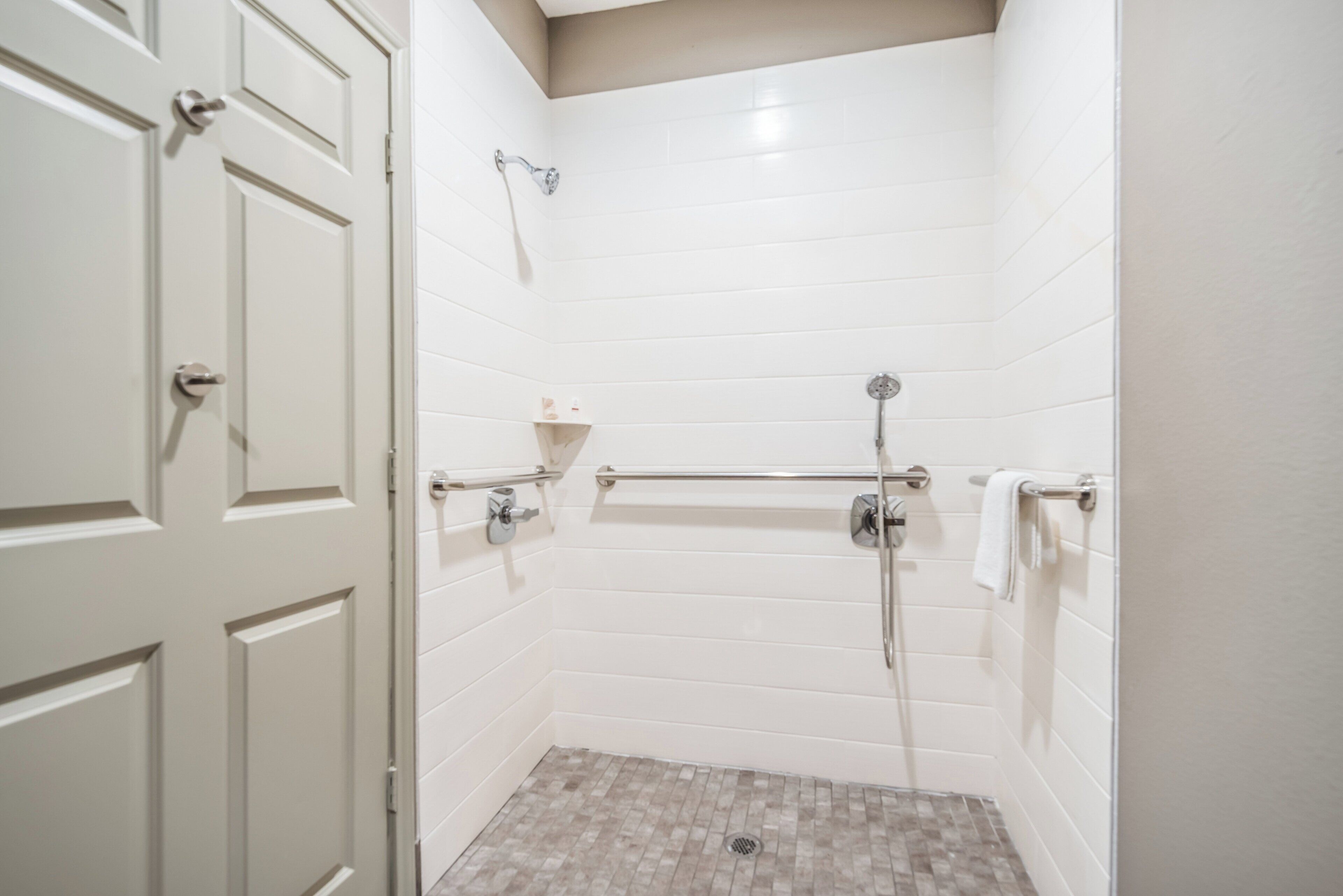 A bathroom with white tiled walls, a shower with a handheld showerhead, a grab bar, and a towel rack.