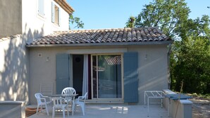 Et lille hus med tegltag, skydedør og en terrasse med hvide plastikstole og et bord.
