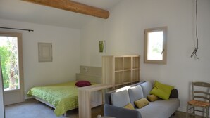 Et lille, godt oplyst værelse med en seng, en sofa, en stol og et skrivebord. Der er vinduer med naturligt lys og udsigt til grønne områder udenfor.