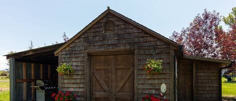 A rustic barn-style building with a large wooden door, two flower baskets on either side, and a covered area to the left.