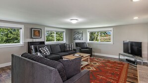 Ein modernes Wohnzimmer mit grauen Sofas, einem Couchtisch und einem Fernseher. Große Fenster und ein Teppich mit rotem Muster sind ebenfalls vorhanden.
