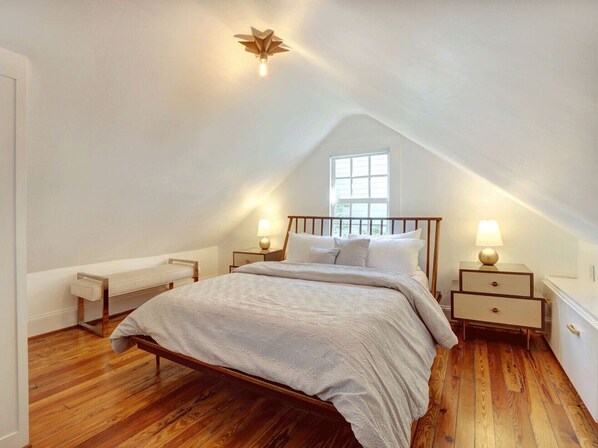 Una camera da letto con soffitto spiovente, un letto con struttura in legno, due comodini con lampade, una panca e una finestra.