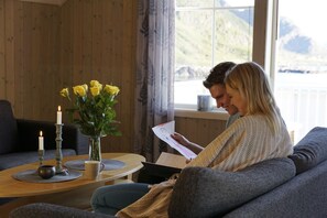 Une pièce chaleureuse au décor intérieur en bois, une grande fenêtre offrant une vue sur les montagnes, un canapé, une table basse avec des bougies et un vase rempli de fleurs jaunes.