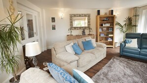 Ein Wohnzimmer mit einem beige farbenen Sofa, blauen Kissen, einem blauen Ledersessel, einem Bücherregal, einem Spiegel und einer Topfpflanze.