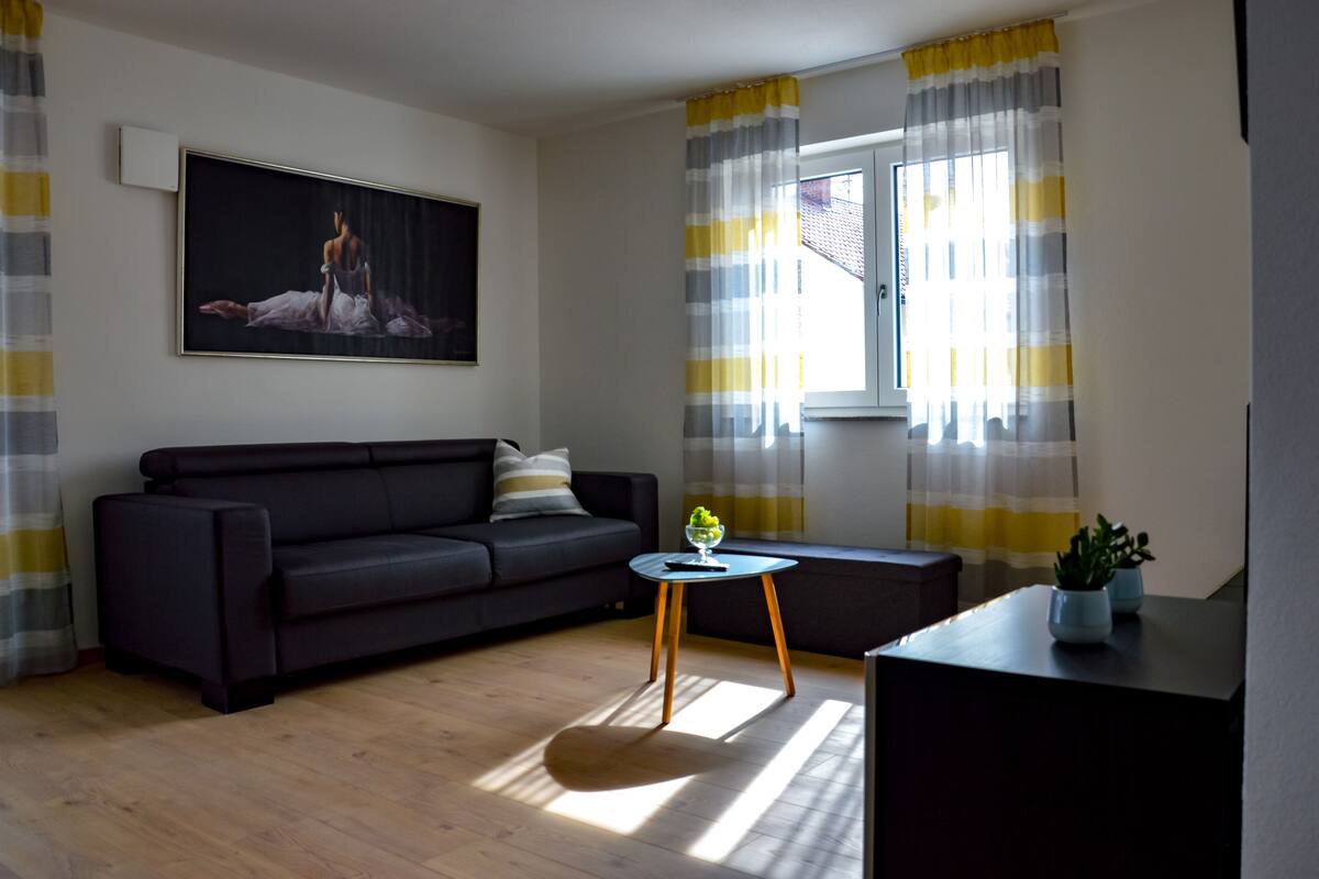 A modern living room with a dark sofa, a small round table with a plant, a painting on the wall, and a window with curtains.