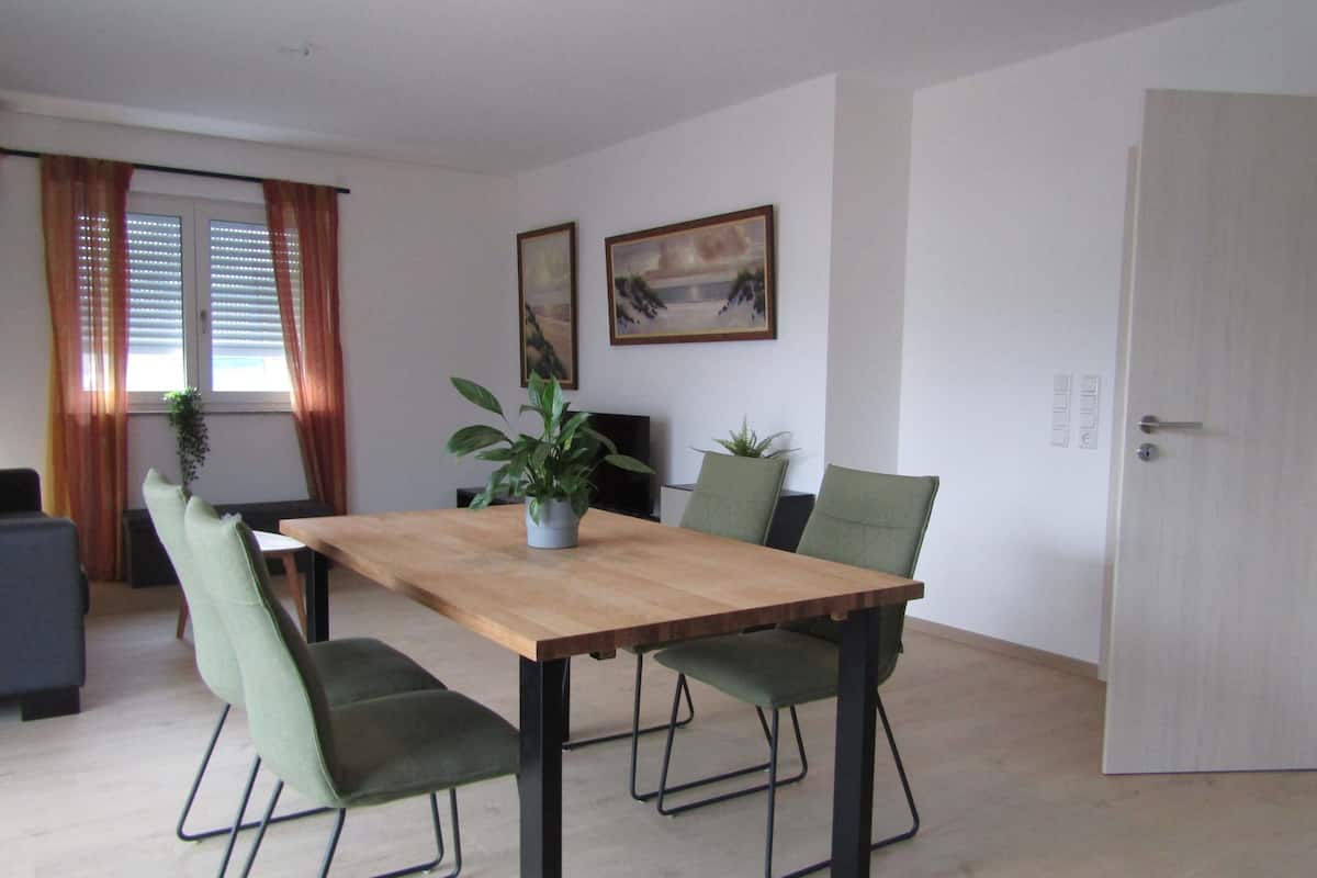 A dining area with a wooden table, four green upholstered chairs, a potted plant, and two framed pictures on the wall.