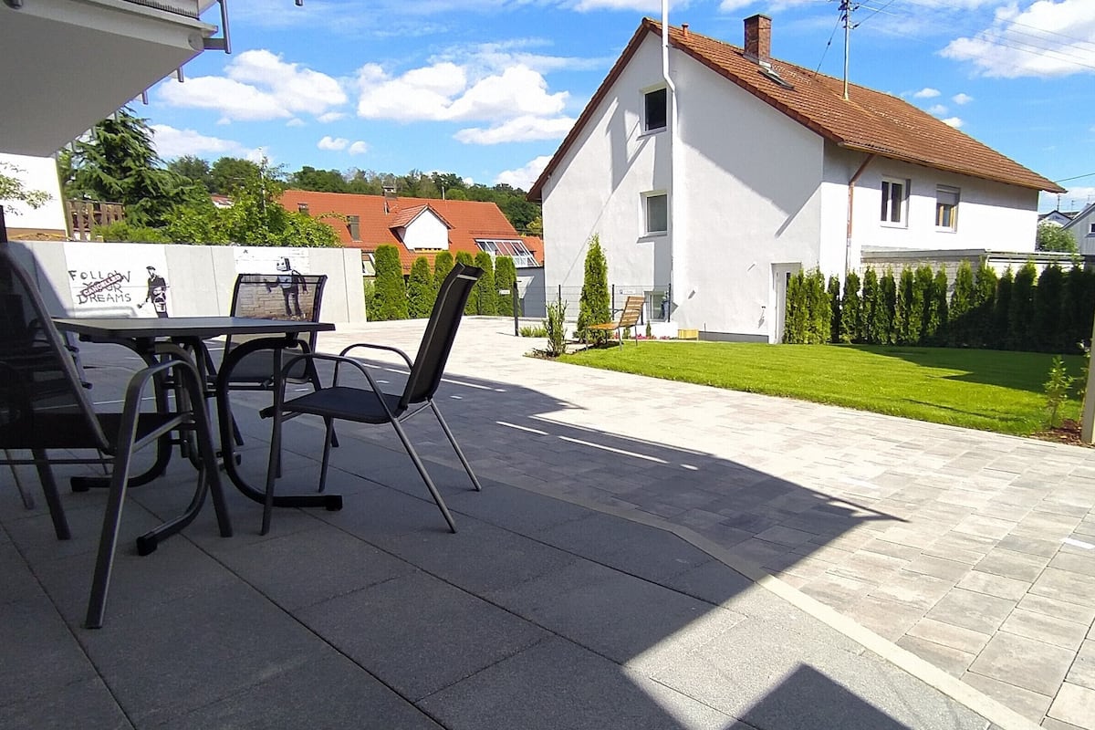 An outdoor patio with a table and chairs, a white house in the background, and a well-maintained lawn.