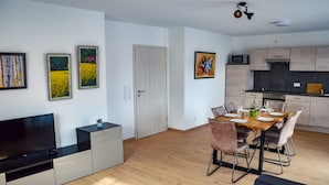 Una sala de estar moderna con televisor de pantalla plana, un comedor con mesa y sillas de madera, y una cocina con armarios de madera clara y un microondas.