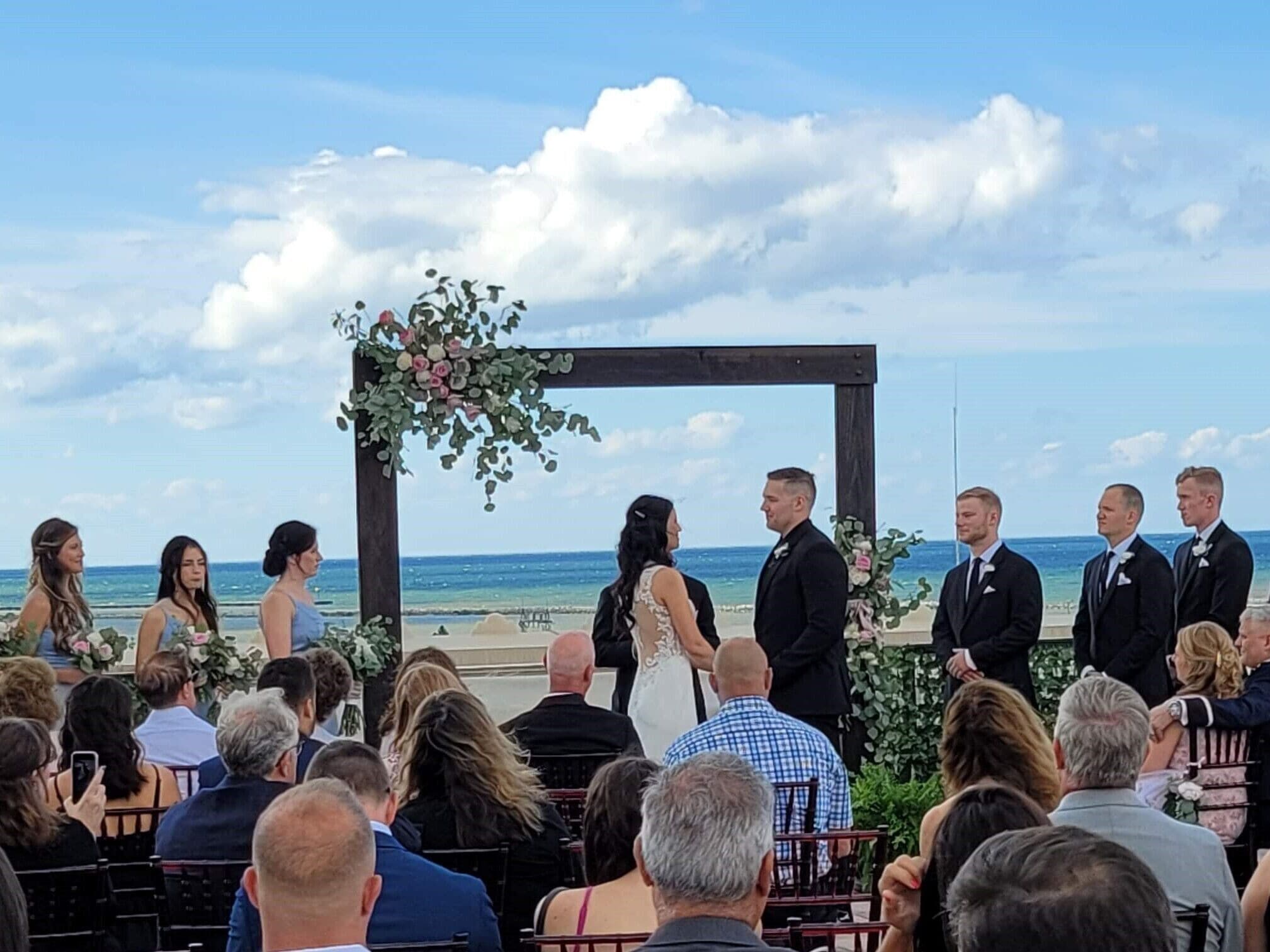 A wedding ceremony outdoors with a wooden arch decorated with flowers, guests seated in rows, and a beach view in the background.