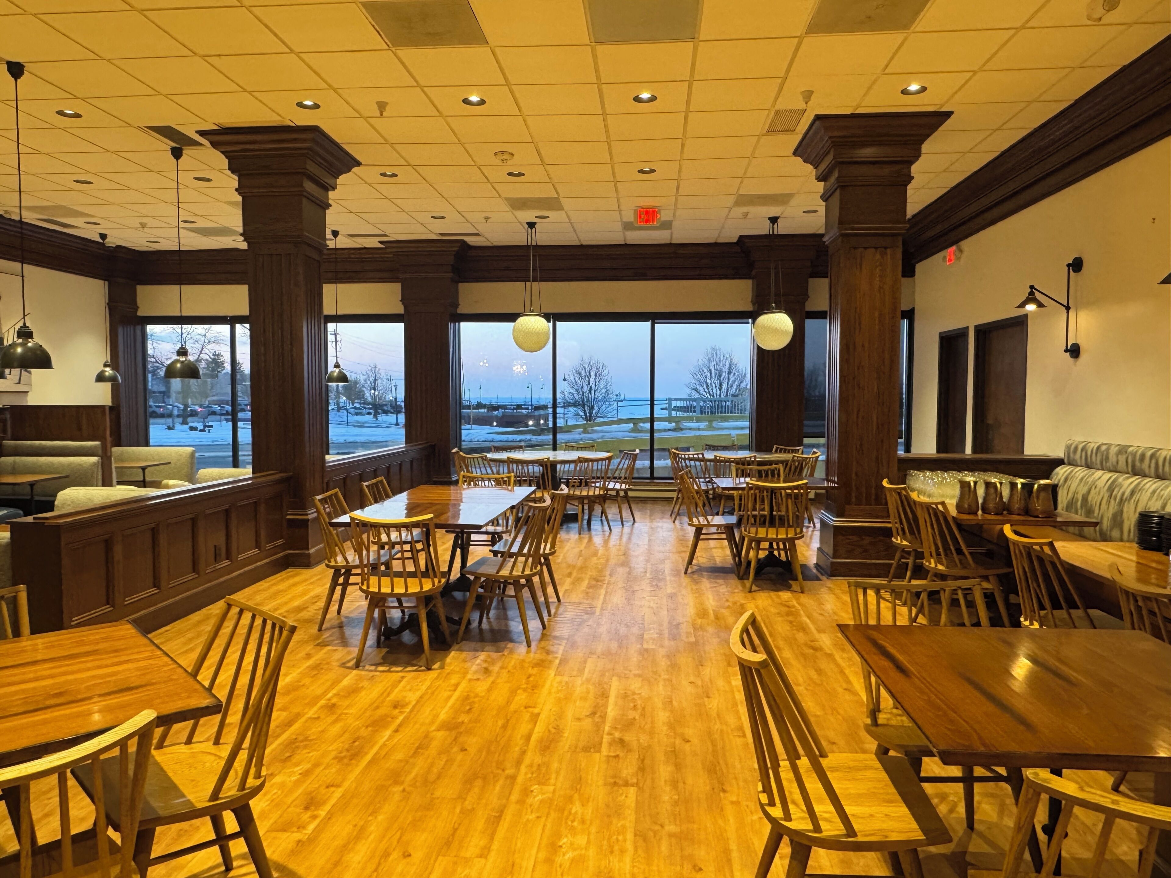 A spacious restaurant with wooden tables and chairs, large windows offering a view of the outdoors, and a high ceiling with recessed lighting.