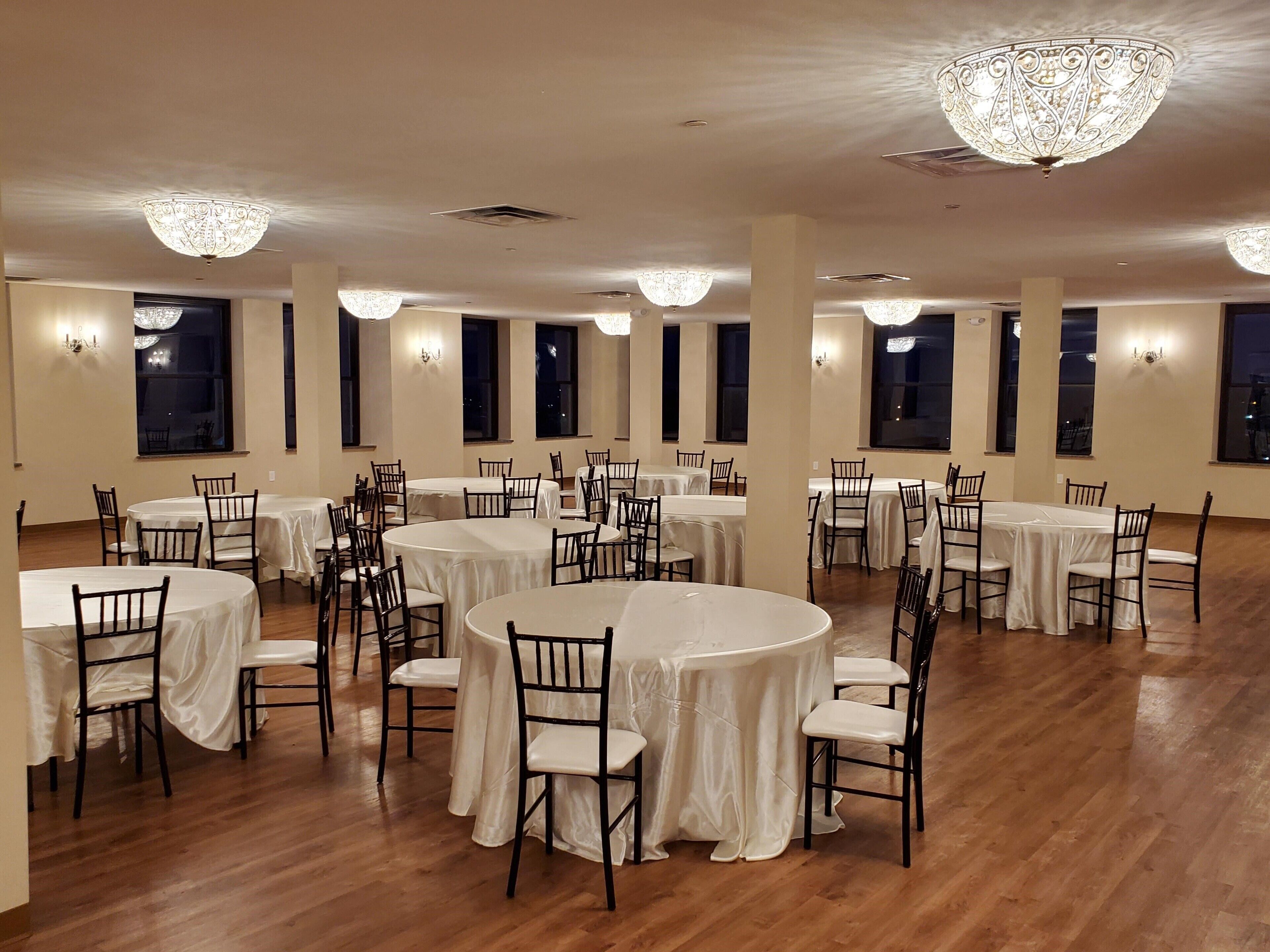 A spacious banquet hall with multiple round tables and chairs, large windows, and decorative chandeliers.