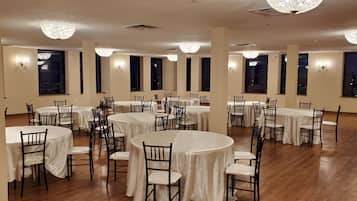 A spacious banquet hall with multiple round tables and chairs, large windows, and decorative chandeliers.