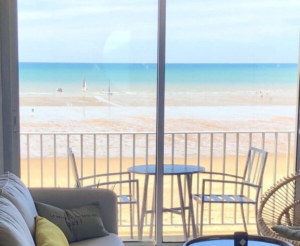 A balcony with a view of a beach, a round table, and a sofa with cushions.