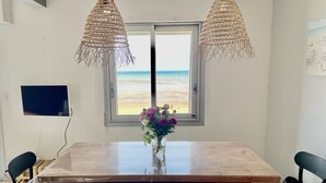 Un comedor con una mesa grande, dos sillas, un jarrón con flores y vistas a la playa a través de una ventana.