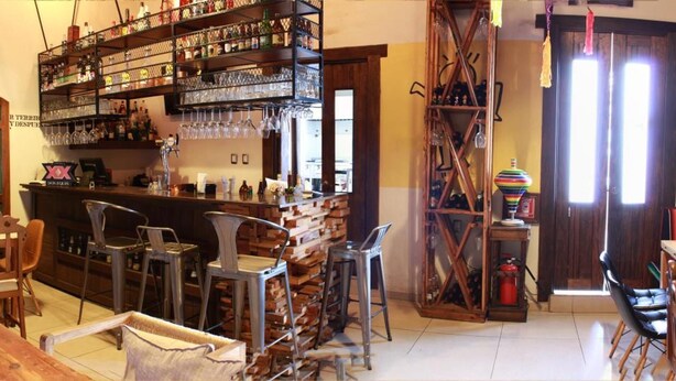 Una zona de bar con mostrador de madera, sillas metálicas y estanterías llenas de botellas y vasos.
